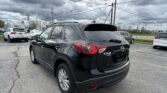 Close-up of a black Mazda CX-5 AWD parked in a car lot with other vehicles and an overcast sky.