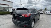 Rear view of a black Mazda SUV (CX-5) in a dealership parking lot, with other cars and a white building nearby on a cloudy day.