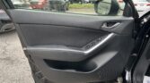 Interior of a black car door panel with power window controls and a silver handle, door open to a parking lot of trucks outside