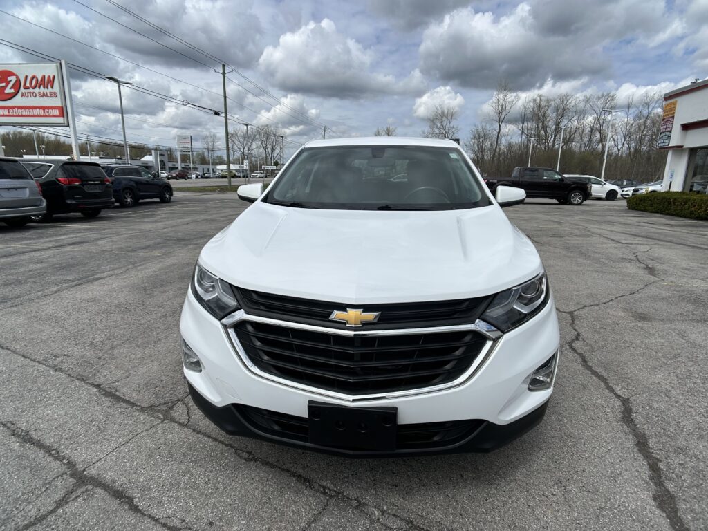 Front view of a white Chevrolet SUV at a car dealership lot, with other vehicles and a cloudy sky behind.