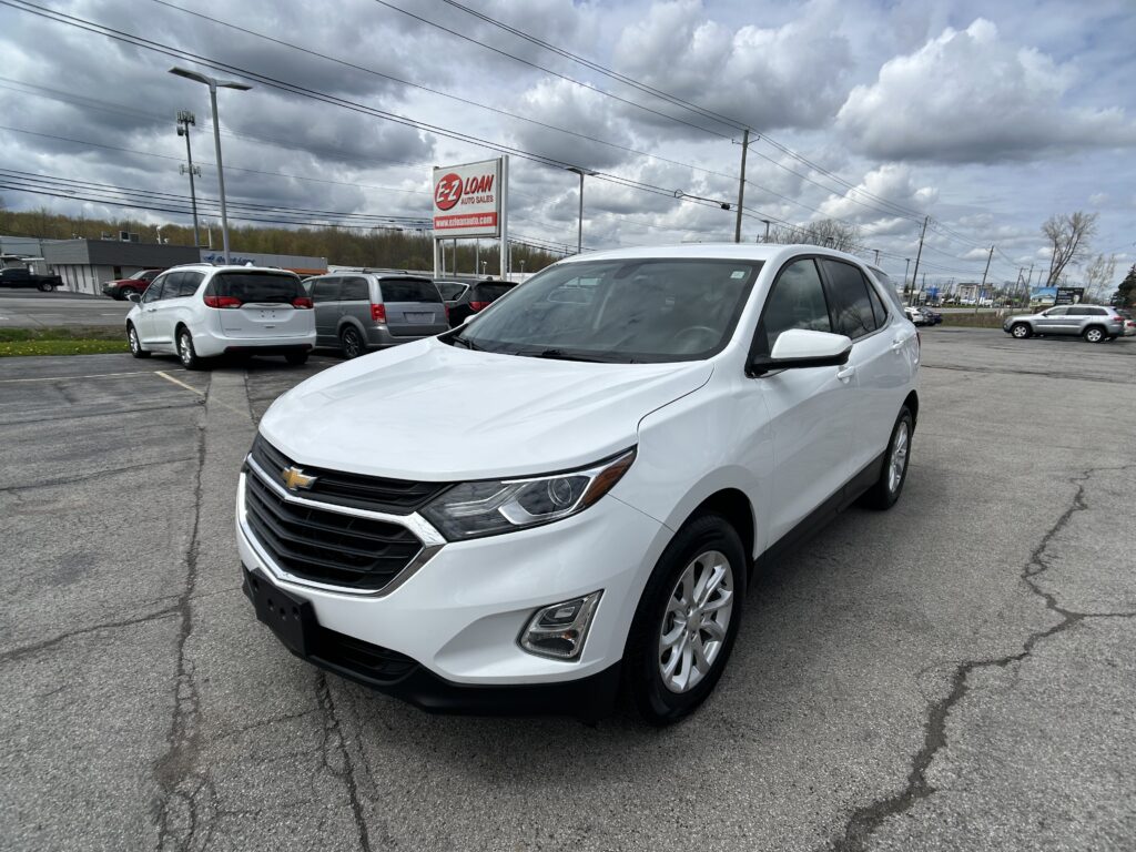 White Chevrolet SUV in foreground at a car dealership lot with other vehicles lined up behind and an EZ Loan Auto Sales sign visible in the background under a cloudy sky, pavement with cracks in the foreground.