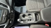 Front interior of a car: center console with gear lever, climate controls, and keys resting in the cup holder; passenger seat nearby and a white sheet on the floor mat.