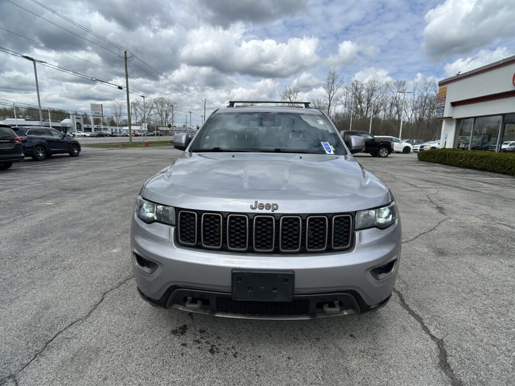 Front view of a silver Jeep SUV parked in a car lot under a cloudy sky with other vehicles in the background