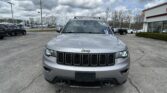 Front view of a silver Jeep SUV parked in a car lot under a cloudy sky with other vehicles in the background