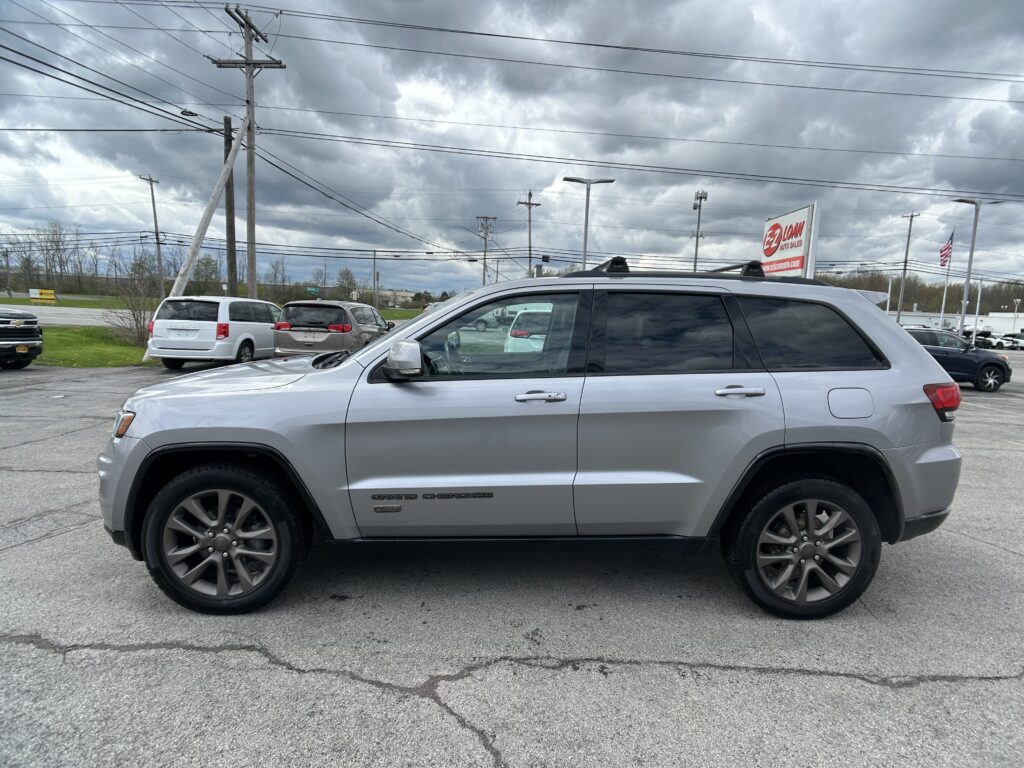 Silver Jeep Grand Cherokee SUV parked in a dealership lot with other vehicles and a cloudy sky above. Overhead power lines run across the scene.