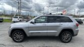 Silver Jeep Grand Cherokee SUV parked in a dealership lot with other vehicles and a cloudy sky above. Overhead power lines run across the scene.
