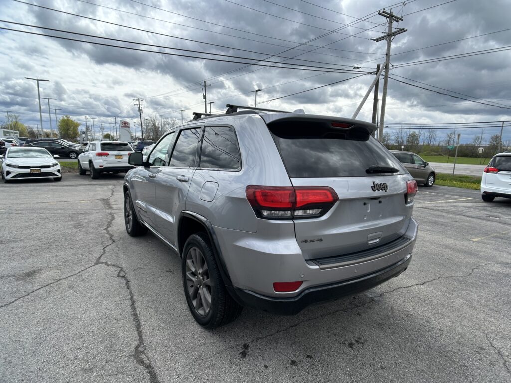 Rear three-quarter view of a silver Jeep SUV in a car dealership lot under a cloudy sky, with other cars nearby.