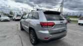Rear three-quarter view of a silver Jeep SUV in a car dealership lot under a cloudy sky, with other cars nearby.