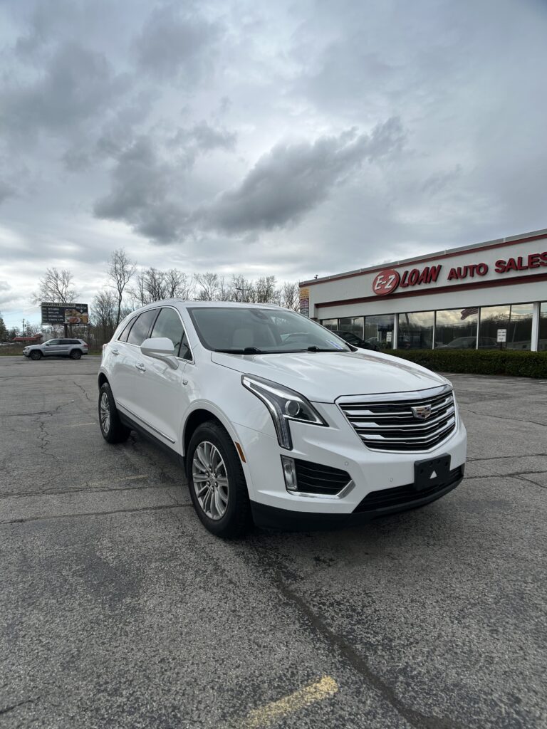 White Cadillac SUV parked in a dealership lot in front of an EZ Loan Auto Sales building with cloudy sky above.