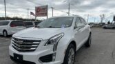 White Cadillac SUV parked in a car lot with an EZ Loan Auto Sales sign and an American flag in the background on a cloudy day,