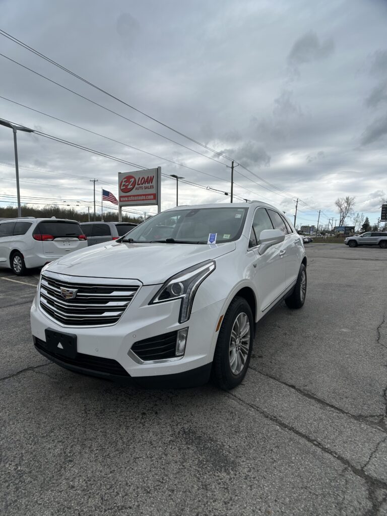 White Cadillac SUV parked in a car lot with an EZ Loan Auto Sales sign and an American flag in the background on a cloudy day,