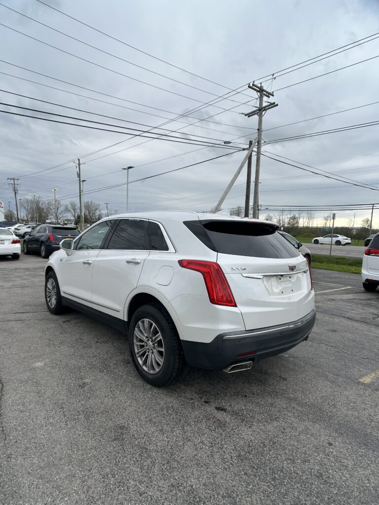 Rear three-quarter view of a white Cadillac XT5 SUV parked in a lot with a cloudy sky and utility poles in the background.
