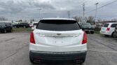 Rear view of a white Cadillac XT5 in a parking lot with other cars and cloudy skies above, power lines crossing the scene.