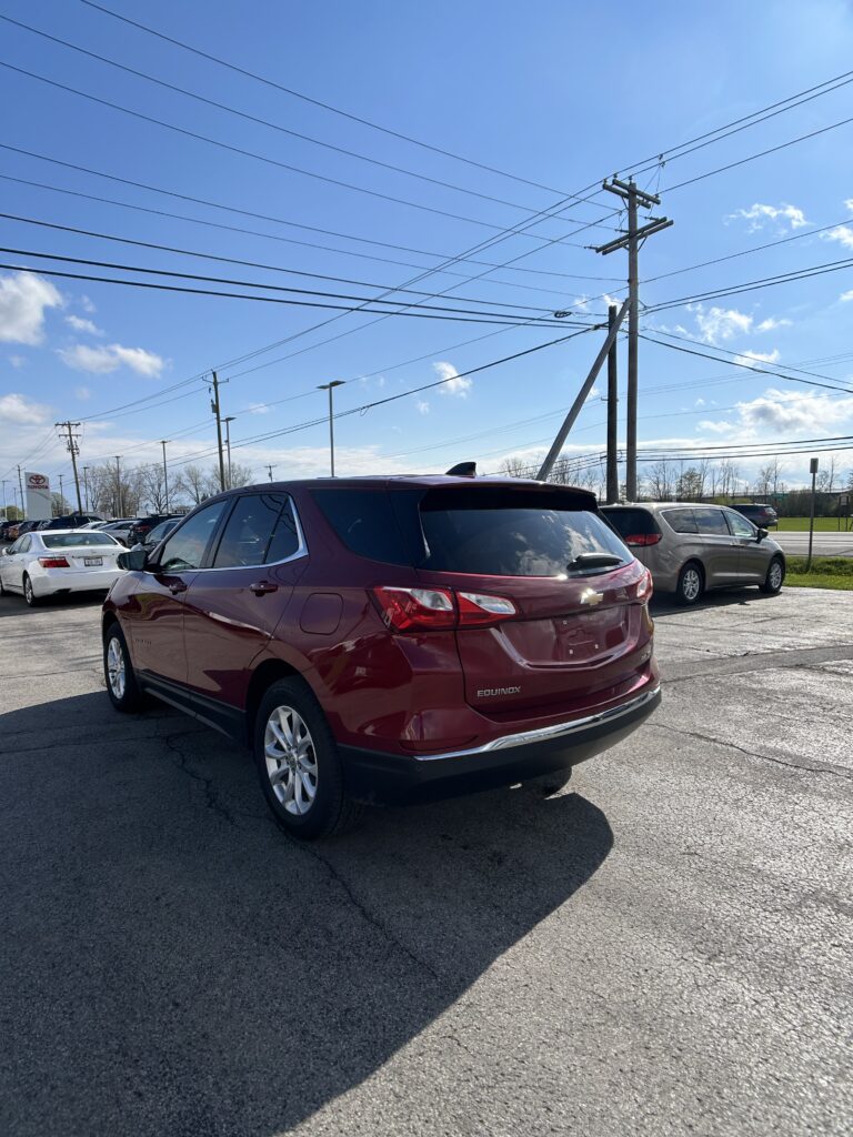 Rear three-quarter view of a red Chevrolet Equinox parked in a dealership lot with power lines overhead and a blue sky.