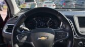 Driver's perspective from inside a Chevrolet, steering wheel centered with emblem visible, dashboard gauges in view, parked in a crowded lot; blue sky and power lines outside through the windshield with several cars ahead.