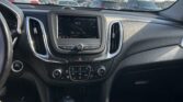 Interior view of a car dashboard from the driver's seat, showing the center touchscreen, climate controls, gear shifter, and cup holders with a parking lot of cars outside through the windshield era of power lines and blue sky.