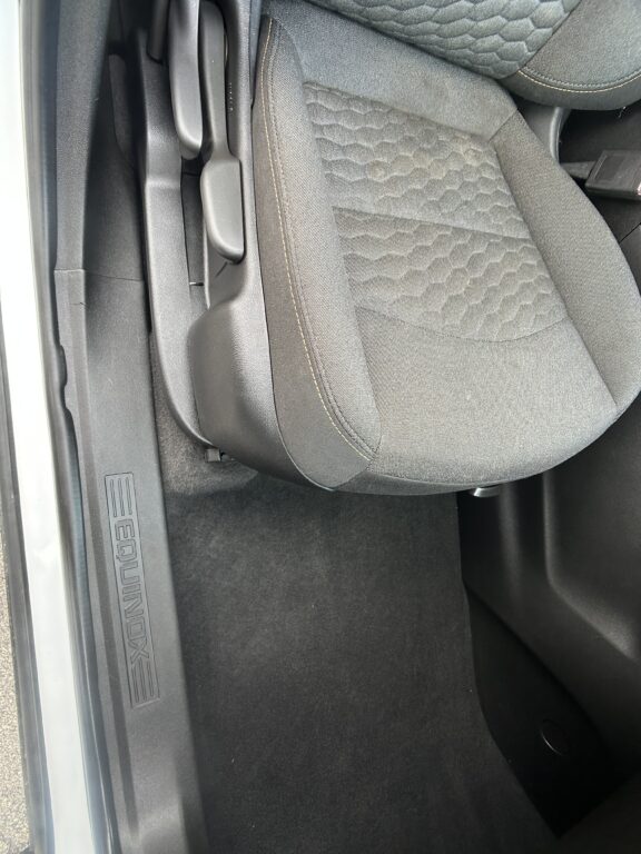 Front passenger seat in a car, black fabric with hexagonal stitch pattern and light stitching, beside the door sill.
