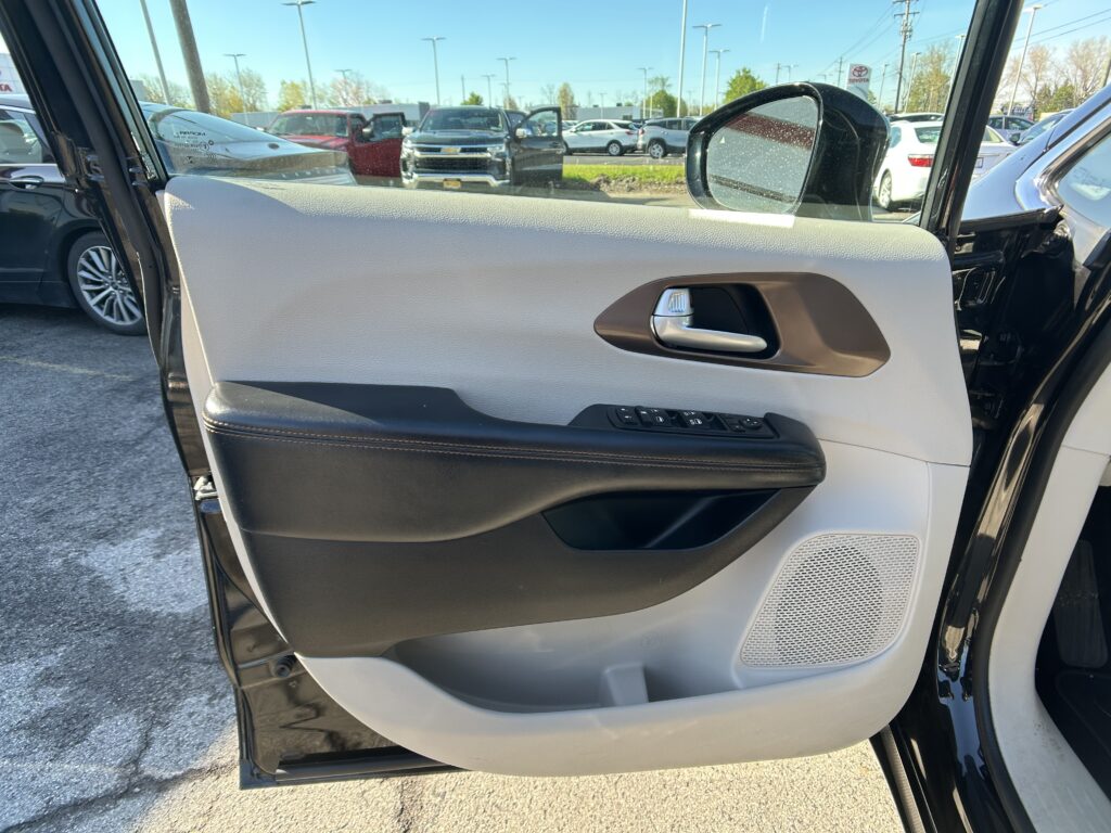 Open car door interior with beige and black trim, window controls, and a view of a parking lot full of cars outside.