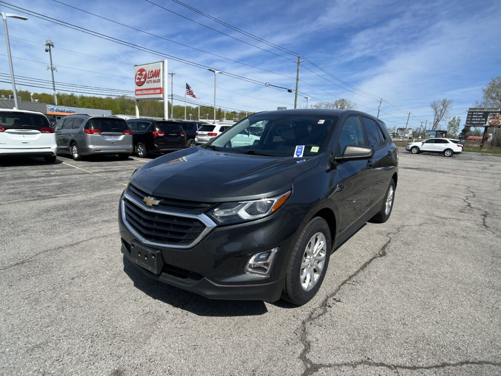 Front-right view of a dark gray Chevrolet SUV in a car dealership lot, with several minivans parked nearby and a blue sky above.