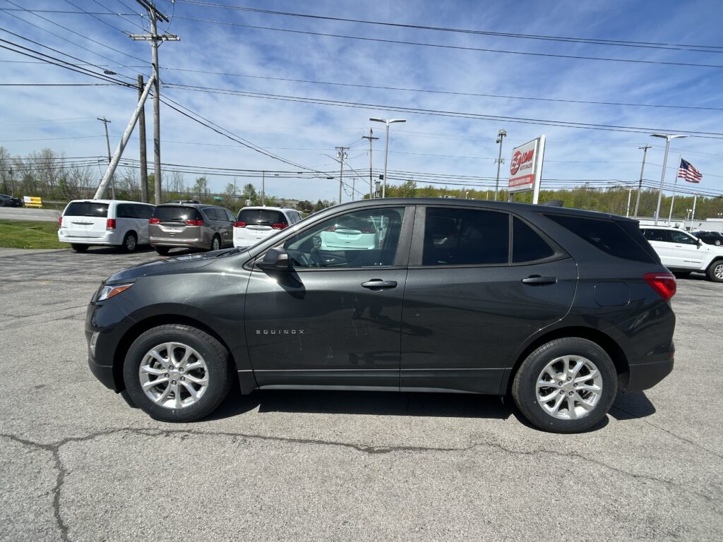 Dark gray Chevrolet Equinox in a car dealership lot, with multiple minivans behind and a dealership sign in the background under a blue sky with power lines overhead.