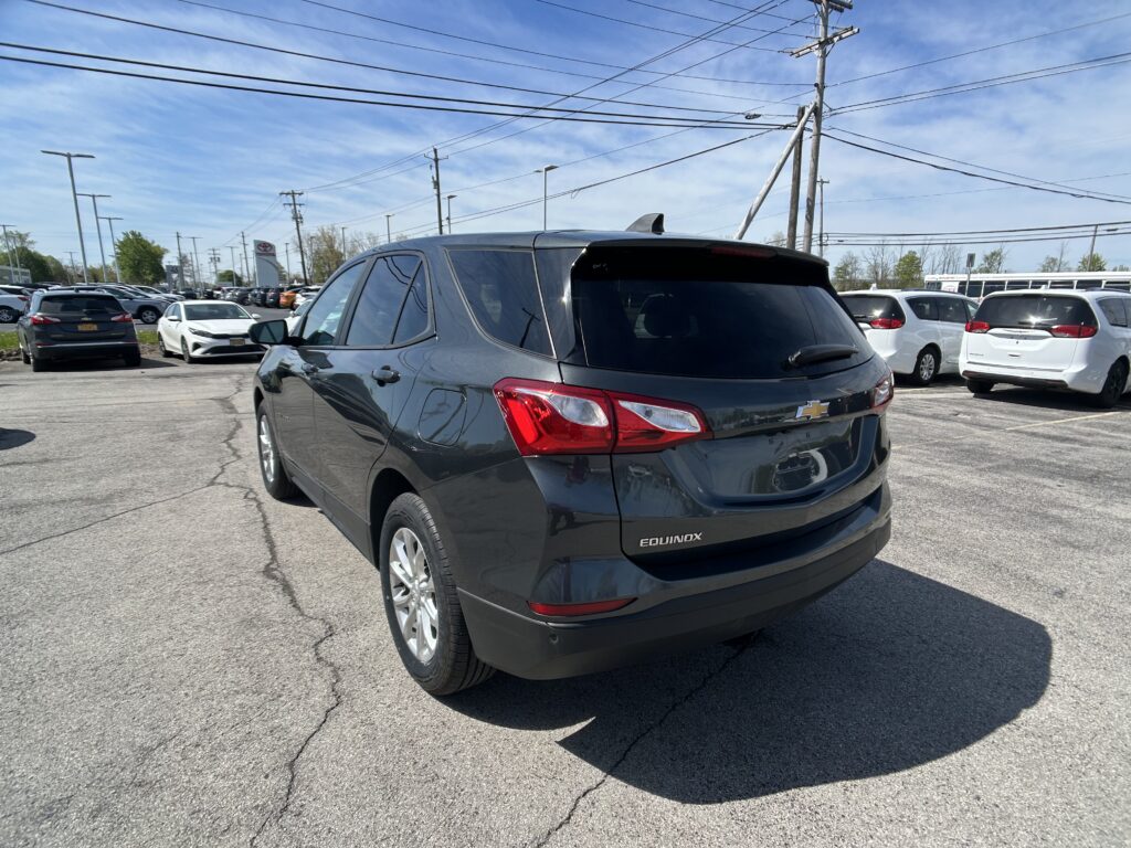 Rear three-quarter view of a gray Chevrolet Equinox SUV in a crowded dealership parking lot under a blue sky with power lines overhead.