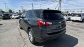 Rear three-quarter view of a gray Chevrolet Equinox SUV in a crowded dealership parking lot under a blue sky with power lines overhead.