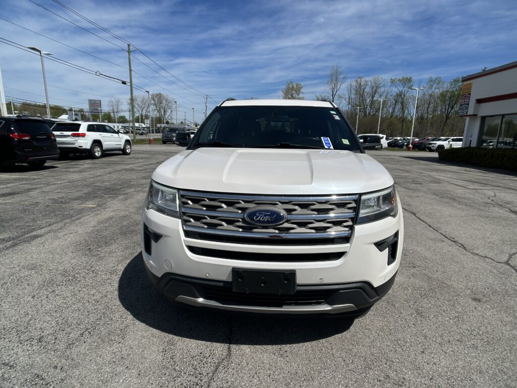 Front view of a white Ford SUV in a dealership lot, with other cars lined up and a building on the right under a clear blue sky.