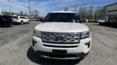 Front view of a white Ford SUV in a dealership lot, with other cars lined up and a building on the right under a clear blue sky.