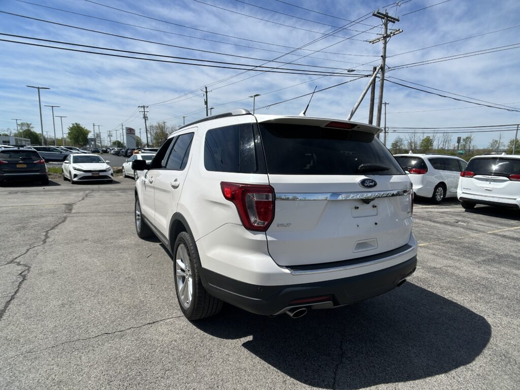 White Ford Explorer XLT seen from the rear-side in a crowded dealership parking lot under a blue sky with overhead power lines above.