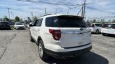 White Ford Explorer XLT seen from the rear-side in a crowded dealership parking lot under a blue sky with overhead power lines above.