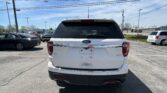 Rear view of a white Ford Explorer SUV in a parking lot, with other cars and overhead power lines against a blue sky.