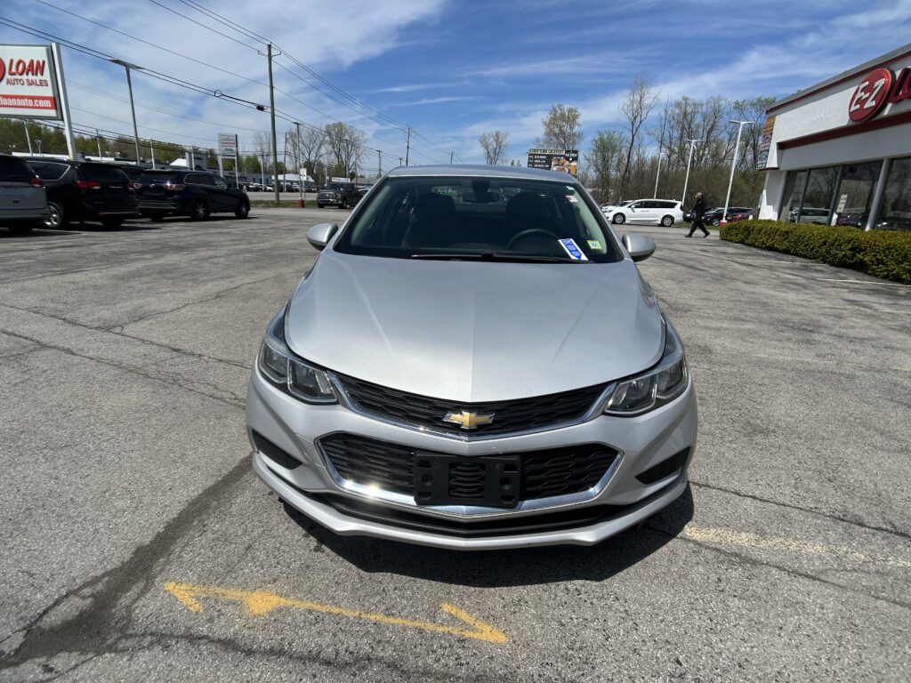 Front view of a silver Chevrolet sedan parked in a car dealership lot with other vehicles and a building in the background.