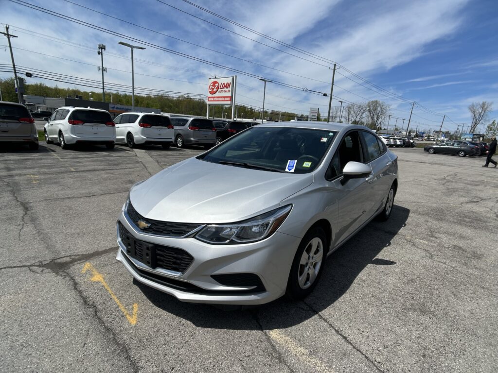 Silver Chevrolet sedan in a parking lot with a row of white vans behind it and a blue sky above.