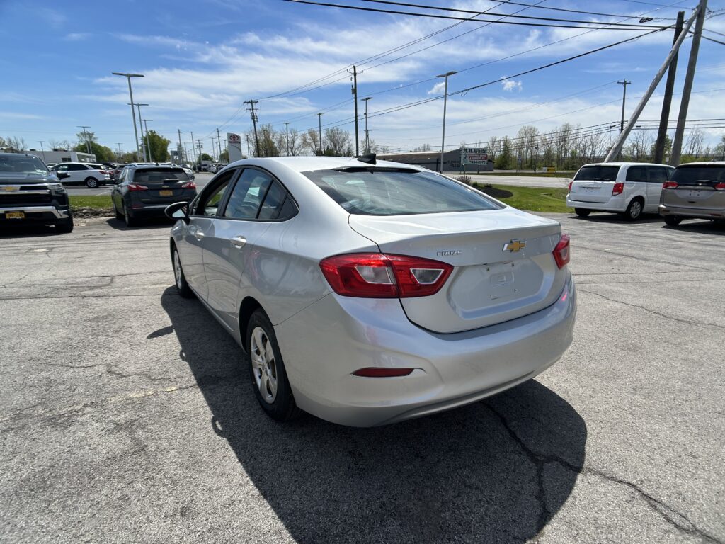 White Chevrolet Cruze rear corner in a busy car dealership lot with other vehicles and utility poles under a blue sky.