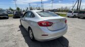 White Chevrolet Cruze rear corner in a busy car dealership lot with other vehicles and utility poles under a blue sky.