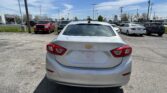 Rear view of a silver Chevrolet Cruze in a parking lot at a car dealership, with other vehicles and dealership buildings in the background on a sunny day.