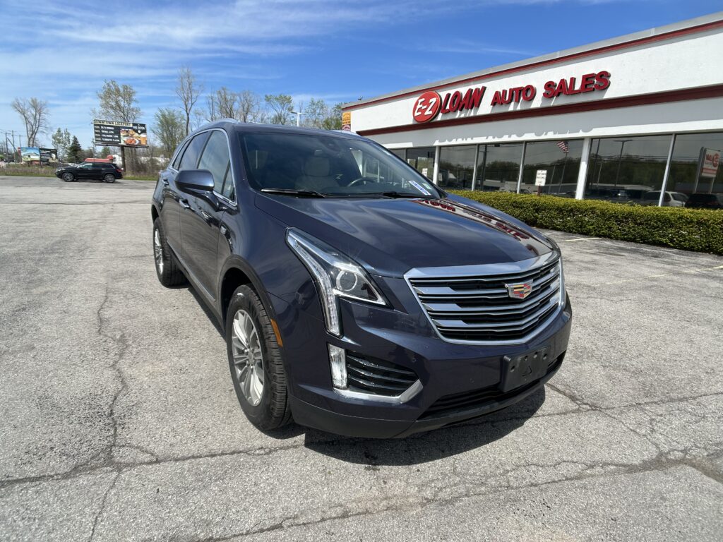 Dark blue Cadillac SUV in the foreground parking lot, in front of an EZ LOAN Auto Sales storefront on a sunny day.