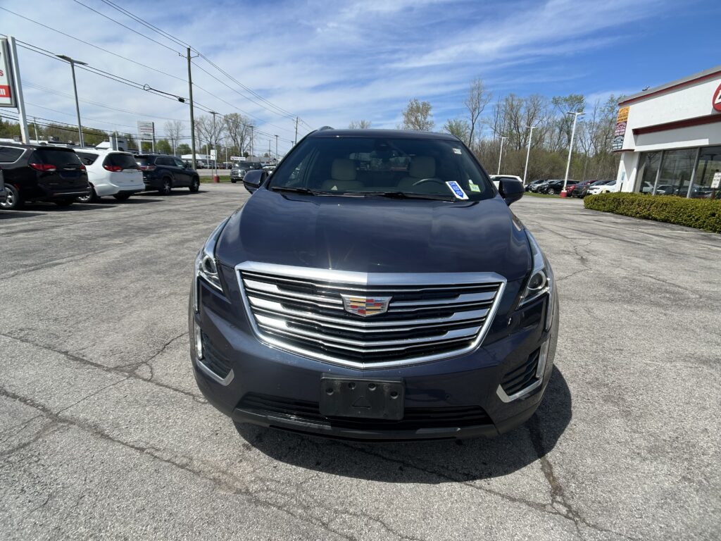 Front view of a dark gray Cadillac SUV in a dealership lot, with other cars and a showroom building on the right.
