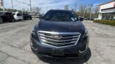 Front view of a dark gray Cadillac SUV in a dealership lot, with other cars and a showroom building on the right.