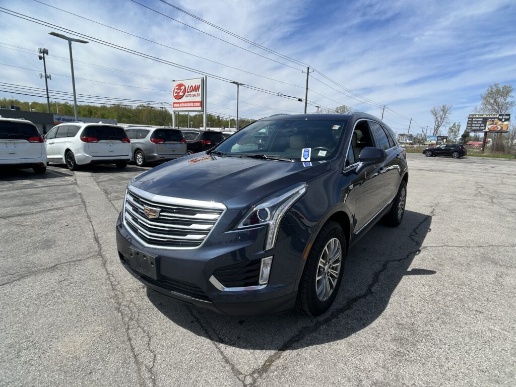 Close-up of a blue Cadillac SUV in a car dealership lot with white minivans in the background and an EZ Loan sign nearby.