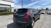 Rear three-quarter view of a dark blue Cadillac SUV in a dealership parking lot with a white building on the left.
