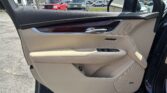 Front passenger door panel of a beige car with wood trim, chrome door handle, and control buttons visible near the armrest; background shows a car dealership lot through the window.