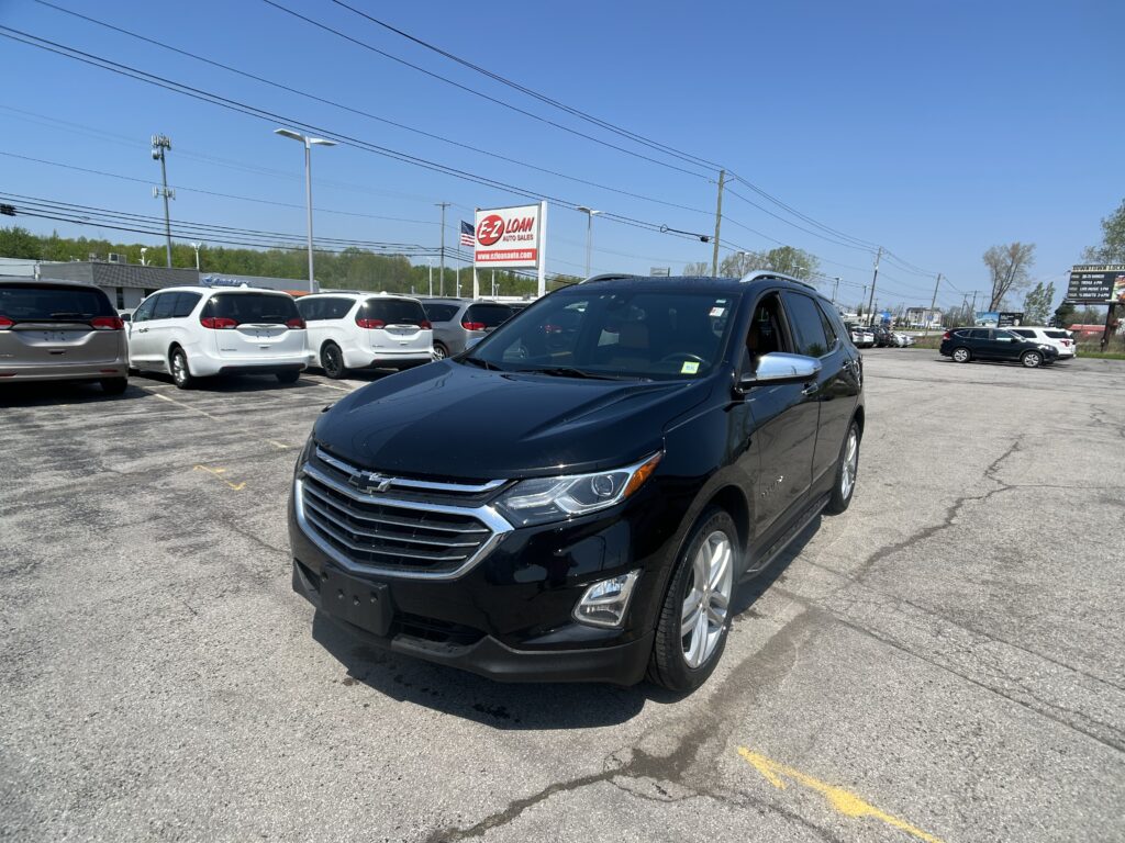 Black Chevrolet SUV in the foreground at a car dealership, with white minivans in a row behind it and an EZ LOAN Auto Sales sign in the distance against a clear blue sky.
