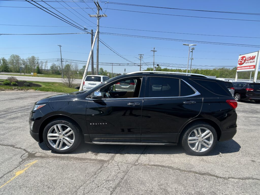 Black Chevrolet Equinox SUV parked in a dealership lot with power lines and utility poles overhead; sign for EZLOAN Auto Sales visible in the background.