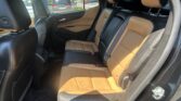 Rear passenger seat area of a car with tan and black leather upholstery, headrests, and visible seat belts. Outside through the window are parked cars and greenery.