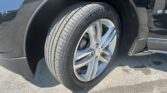 Close-up of a car wheel and tire mounted in a black SUV's wheel well, showing the alloy rim and tread details on the pavement behind it, with the fender visible.