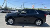 Black SUV in a dealership parking lot with several minivans and power lines overhead, American flag in the distance.