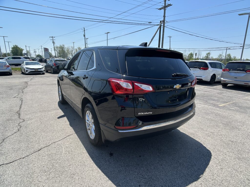 Rear three-quarter view of a black Chevrolet Equinox in a crowded dealership parking lot on a sunny day, with other cars in the background and power lines overhead.
