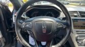 Close-up of a car steering wheel and dashboard with digital gauges and an infotainment screen visible to the right, in a bright showroom lot outside the car