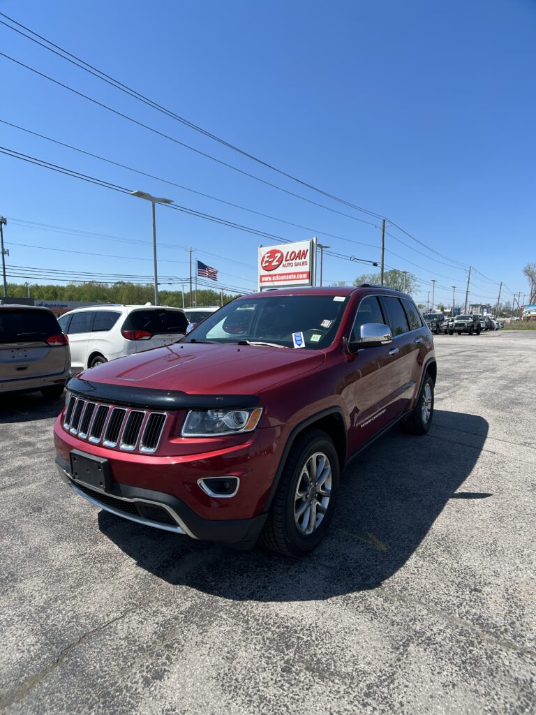 Red SUV in the foreground at an EZ LOAN Auto Sales lot, with other cars, a sign, and an American flag in a bright blue sky.